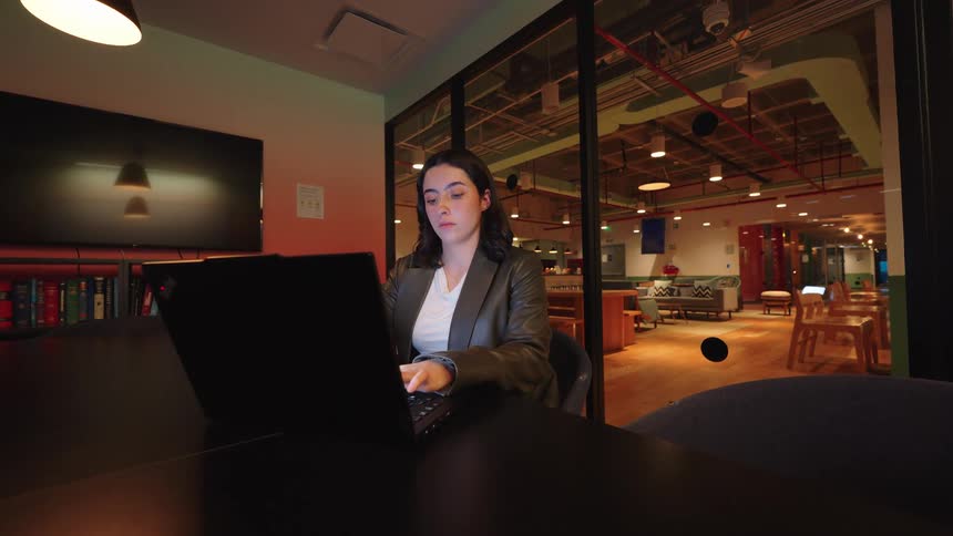 A focused young woman works diligently on her laptop late at night in a modern, glass-walled office space