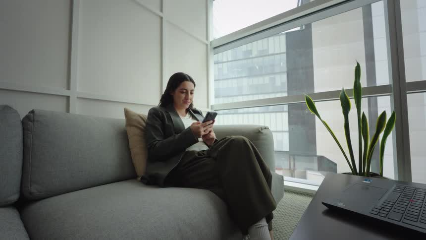 A young businesswoman relaxes on a couch in a modern high-rise office, smiling as she scrolls through her smartphone