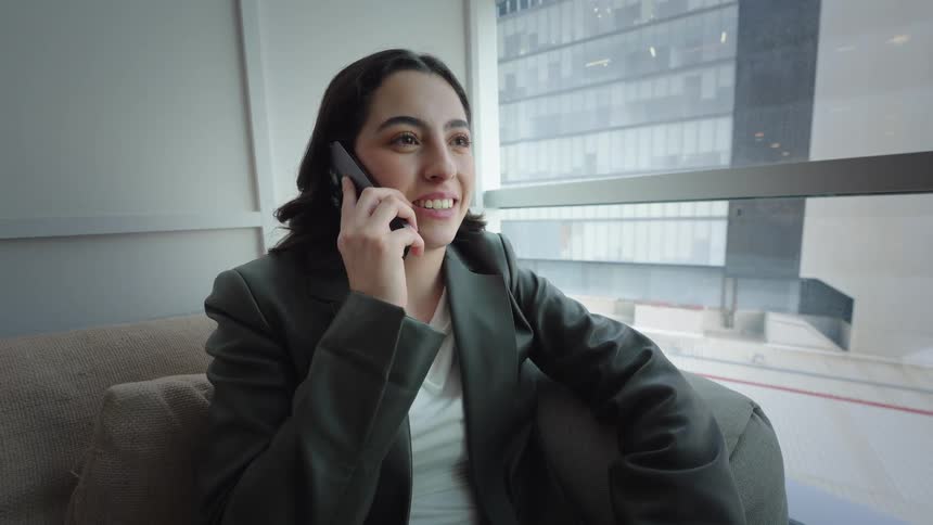 A smiling young businesswoman talks on her smartphone while relaxing on a couch in a modern office with a large window