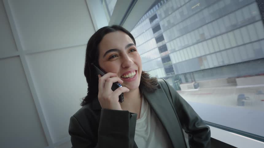 A smiling young professional enjoys a happy phone conversation while standing by a large window in a modern building
