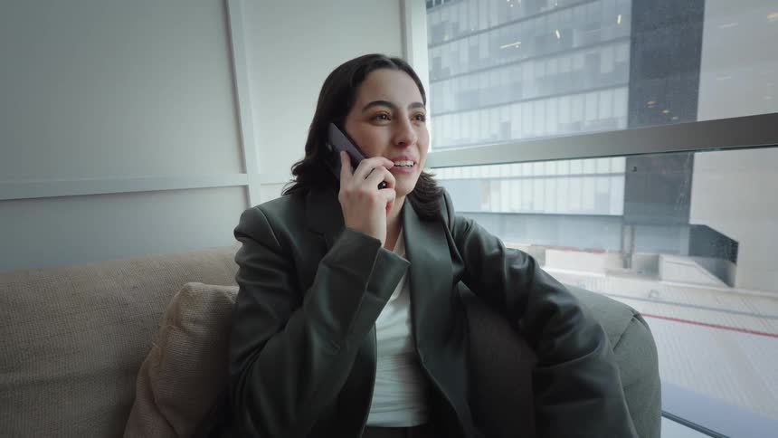 Happy young businesswoman sitting on a sofa in a modern office, enjoying a pleasant conversation on her smartphone by the window