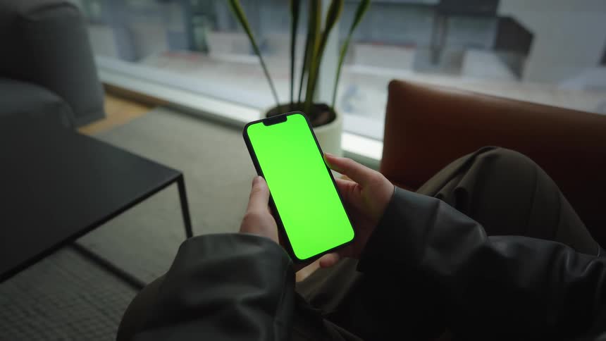 A person relaxes in a modern lounge chair and swipes on a smartphone with a vertical green screen