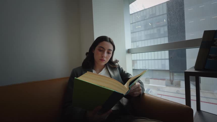A focused young woman in a blazer studies a large book while sitting on a leather couch by a window overlooking the city