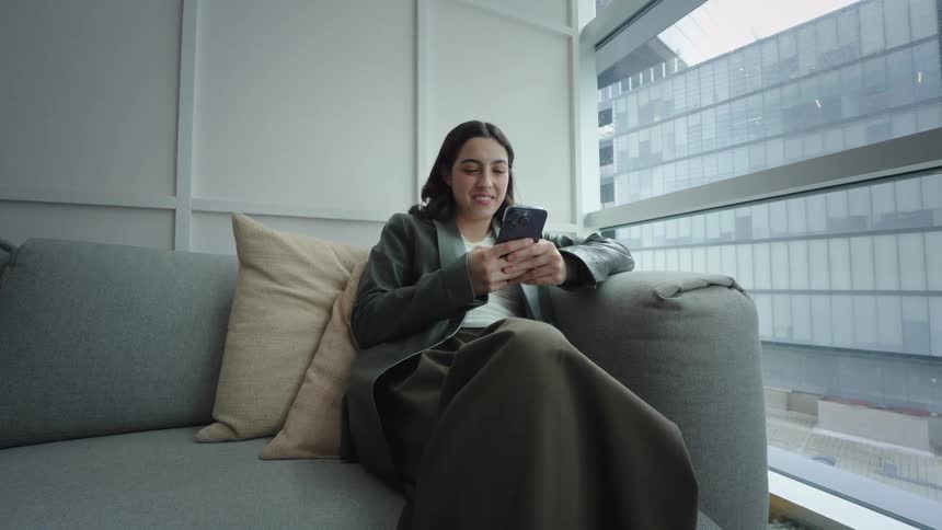 A stylish young woman smiles and laughs while using her smartphone, sitting on a comfortable couch in a modern office space by a large window