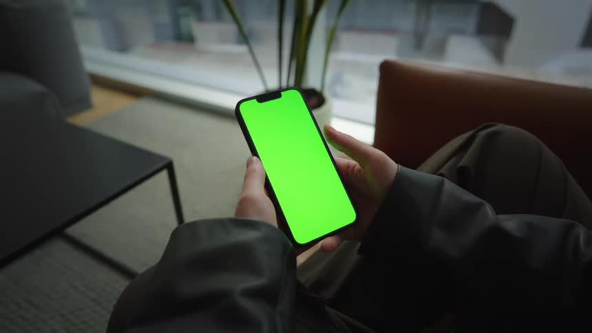 A person scrolls and taps on a smartphone with a green screen, viewed from a first-person perspective in a modern living room
