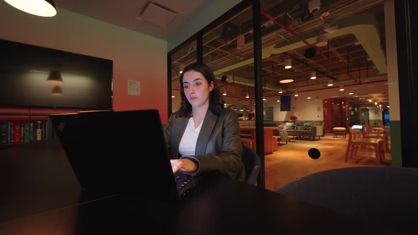 A focused young businesswoman works diligently on her laptop in a modern conference room at night