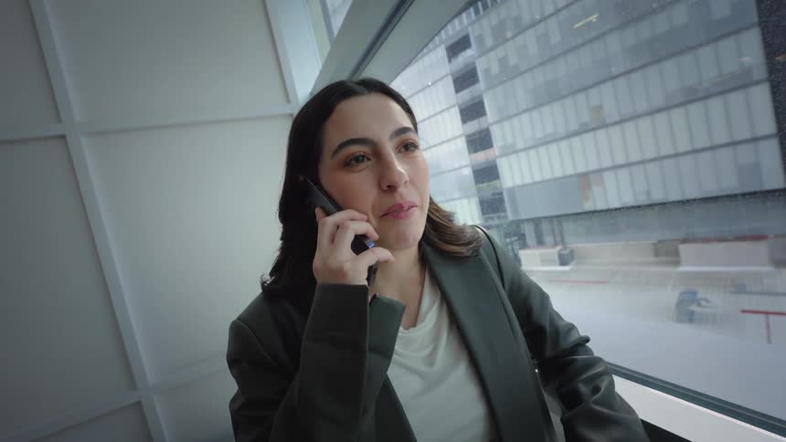 A young businesswoman stands by a window in a modern office, happily answering a call on her smartphone with a city view in the background