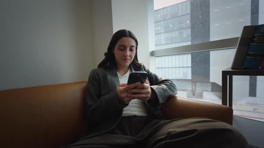 A young businesswoman smiles while using her smartphone, relaxing on a leather couch in a modern high-rise office