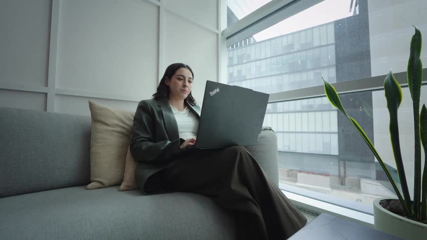 A professional woman sits comfortably on a couch in a modern high-rise office while working on her laptop during a video call