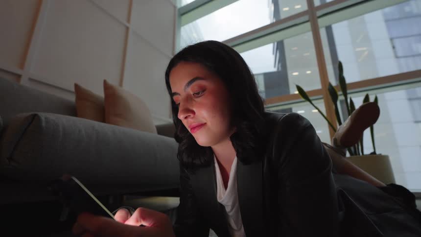 A smiling young businesswoman lies on the floor in a modern high-rise office, happily browsing on her smartphone after hours