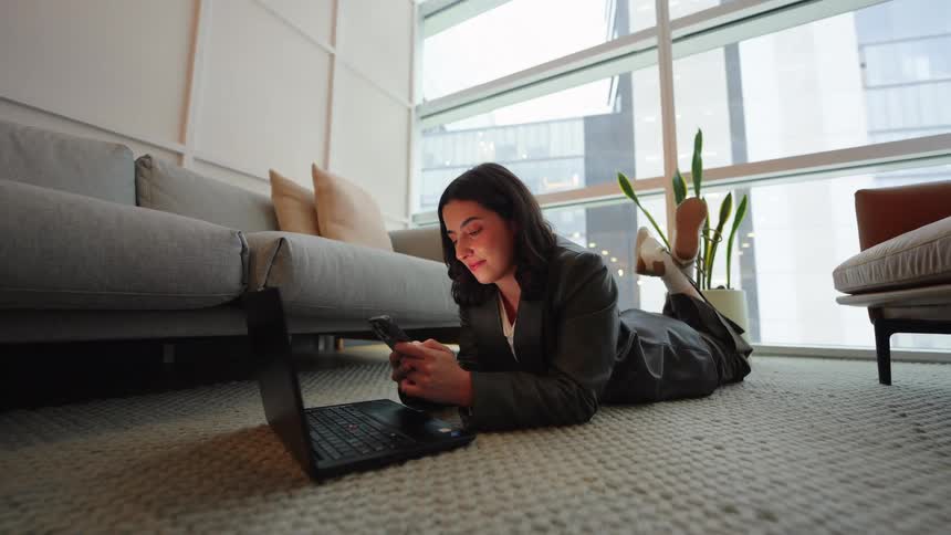 A stylish young woman in a suit lies on the floor of her modern apartment, scrolling on her smartphone with her laptop nearby