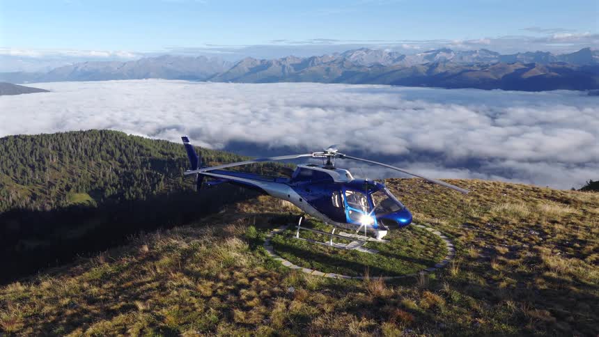 A blue helicopter with spinning rotor blades rests on a mountain helipad high above a sea of clouds and a distant mountain range