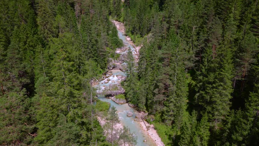 Aerial drone view slowly approaches a charming wooden bridge spanning a rushing turquoise river in a lush green mountain forest
