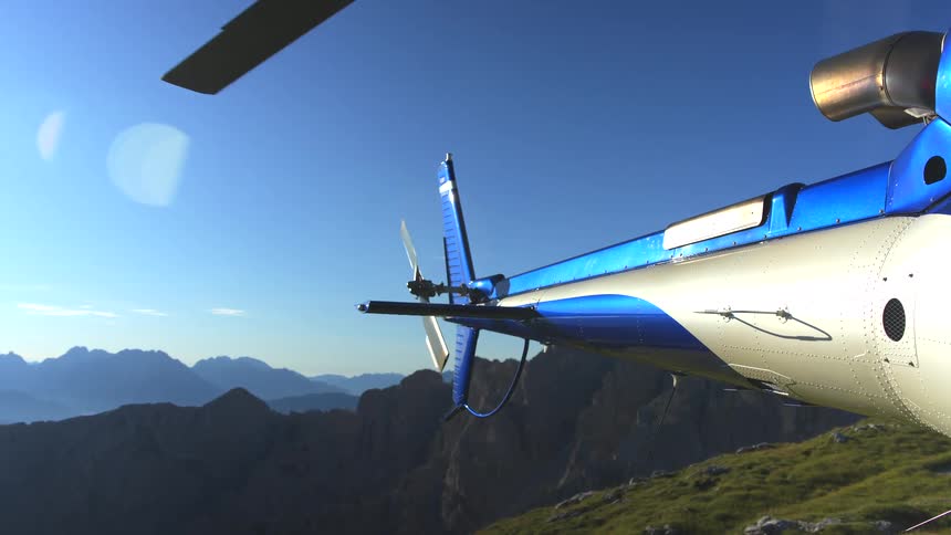 A helicopter's spinning tail rotor is seen up close as it hovers above a dramatic mountain range on a sunny day
