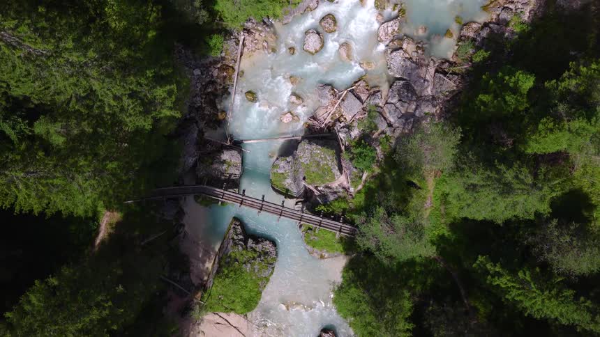 An aerial top-down view follows a stunning turquoise river as it flows through a dense evergreen forest and under a wooden footbridge in the mountains