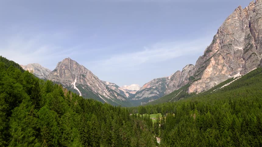 A slow aerial shot pulls back over a vast green forest to reveal a majestic mountain valley on a clear summer day