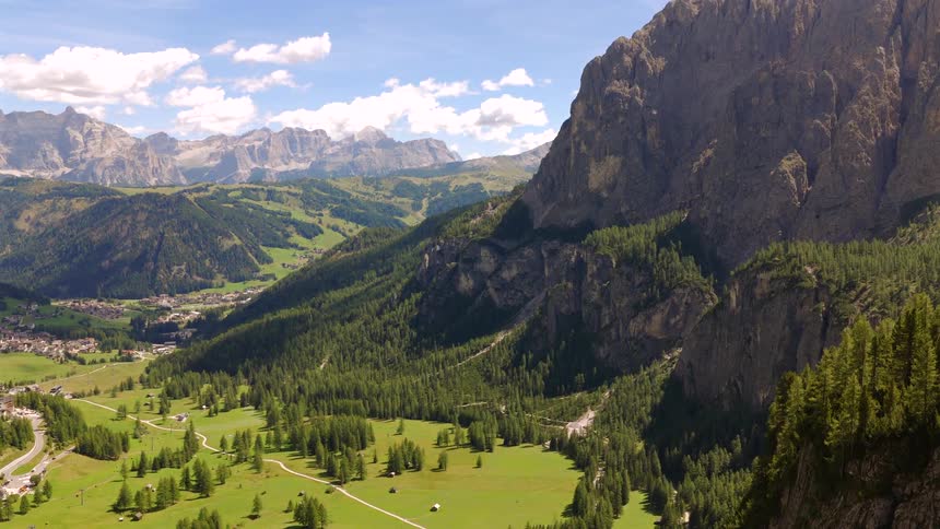Stunning aerial view pulls back to reveal a lush green valley and a distant village nestled in the majestic Dolomite mountains of Italy