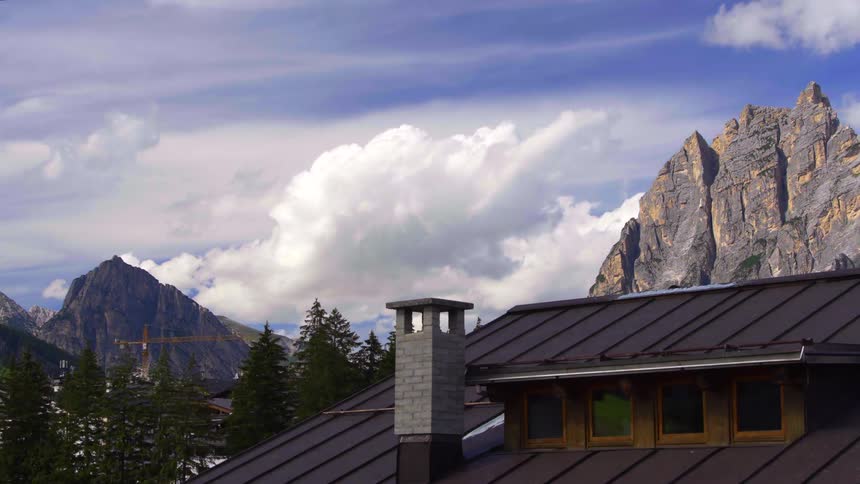 A timelapse of white clouds moving quickly across a blue sky above jagged mountain peaks and a rustic cabin roof