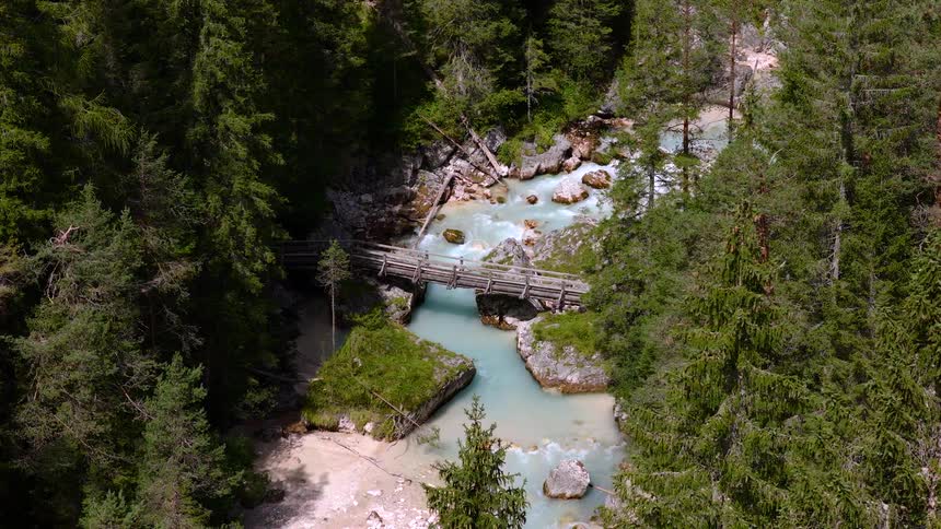 A high-angle drone shot slowly pulls back to reveal a turquoise glacial river flowing under a wooden bridge in a dense evergreen forest