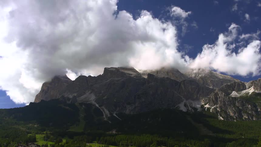 A dramatic time-lapse shows fluffy white clouds rolling over a majestic mountain range with lush green forests below
