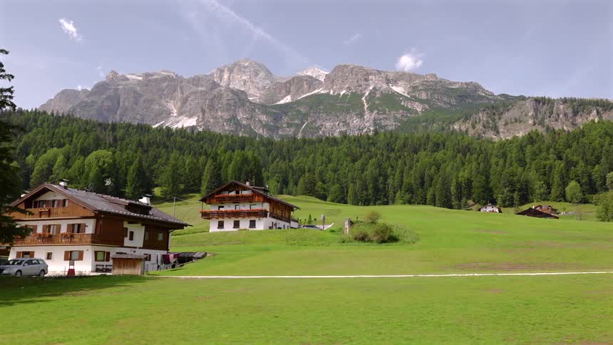 A slow pull-back shot reveals an idyllic mountain landscape with traditional alpine houses nestled in a verdant meadow below the towering peaks of the Dolomites