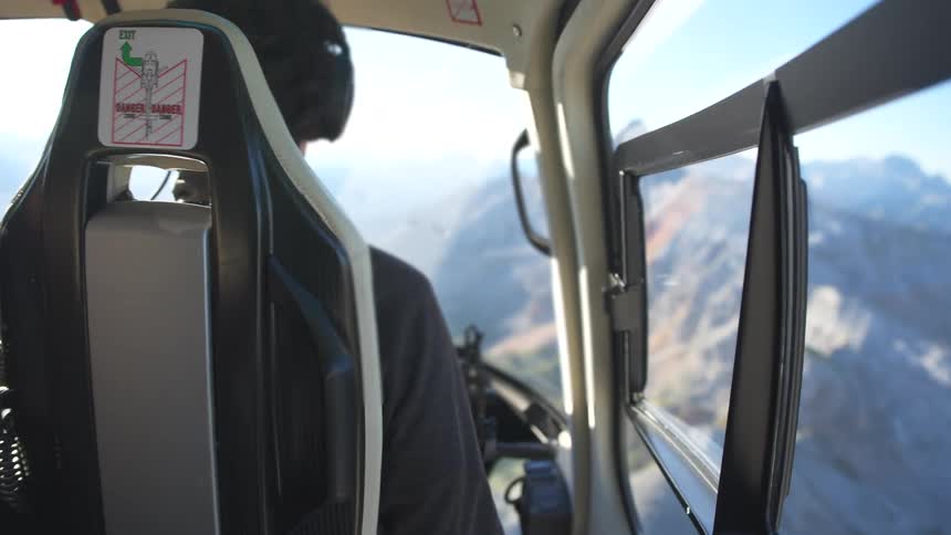 An over-the-shoulder perspective from inside a helicopter reveals a pilot expertly navigating over a breathtaking, sunlit mountain range