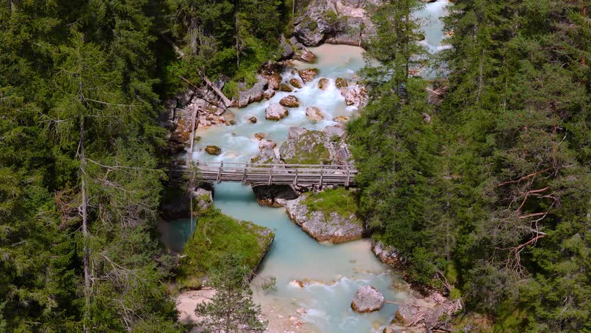 A picturesque wooden footbridge crosses a fast-flowing turquoise mountain river surrounded by a dense pine forest