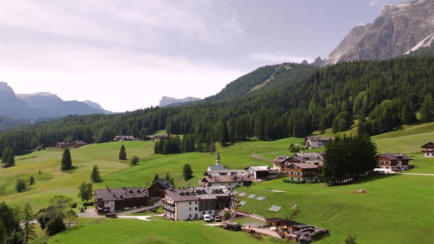 A scenic wide shot of a peaceful village nestled in a lush green valley at the foot of the Italian Dolomites