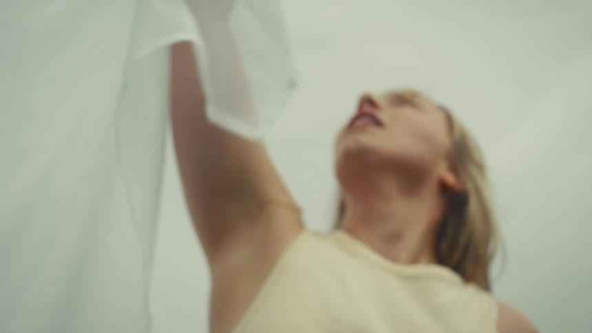 An ethereal slow-motion shot from a low angle reveals a blonde woman behind a sheer white fabric