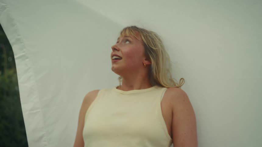 A young blonde woman with an expression of wonder looks up from under a white sheet blowing in the breeze