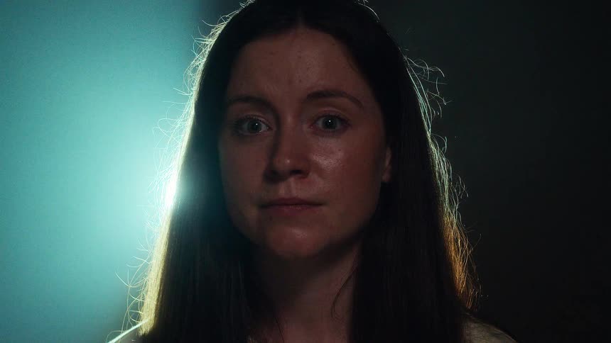 A woman with a serious and stoic expression stares directly at the camera under dramatic, moody lighting