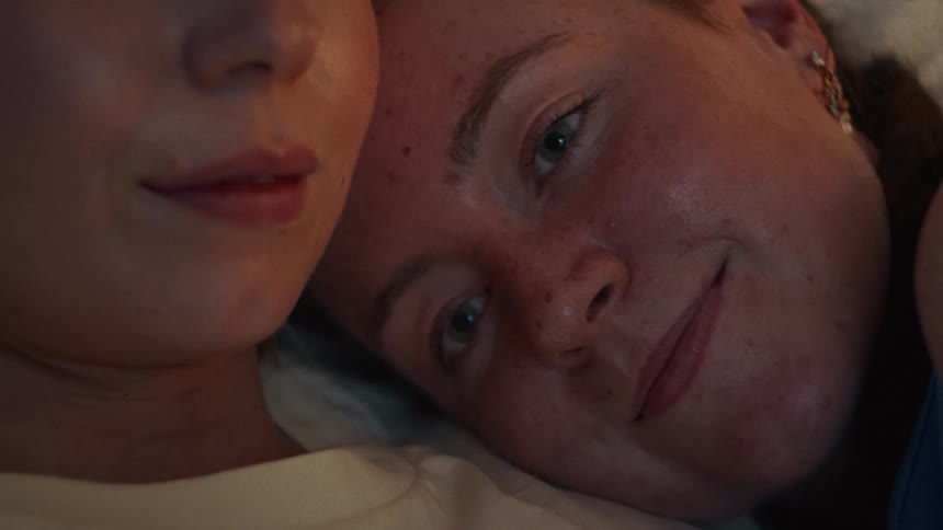 A tender close-up of a young woman with freckles lovingly resting her head on her girlfriend's shoulder in a warm, cozy light