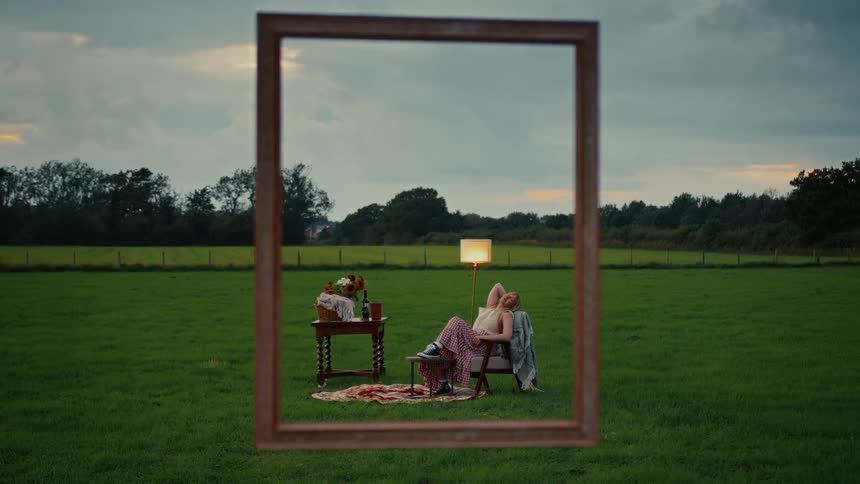 A woman relaxes in an armchair during a surreal picnic in a grassy field at dusk, viewed through an empty picture frame