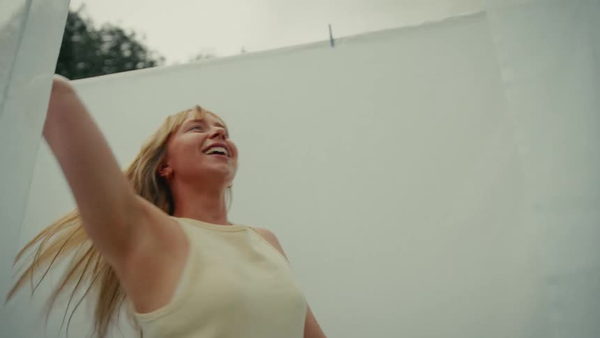 A happy young woman with long blonde hair dances freely among white sheets hanging on a clothesline outdoors