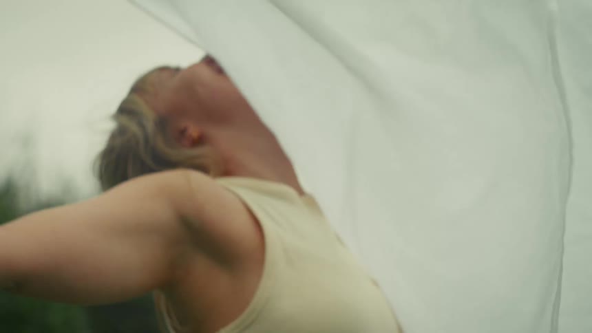 A woman with her head tilted back and arms open wide is slowly covered by a billowing white sheet