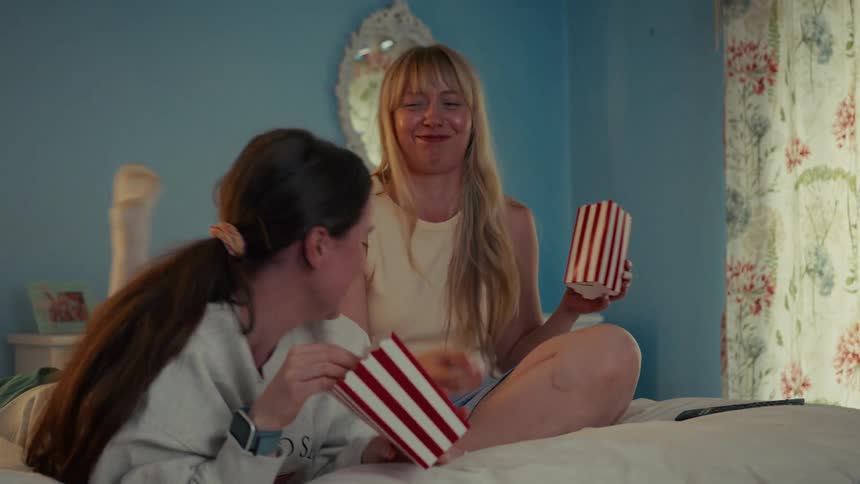 Two playful female friends have a fun movie night in bed, laughing as one tries to catch popcorn thrown by the other in slow motion