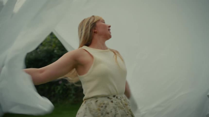 A joyful blonde woman spins in slow motion with a white sheet hanging on a clothesline in a lush green garden