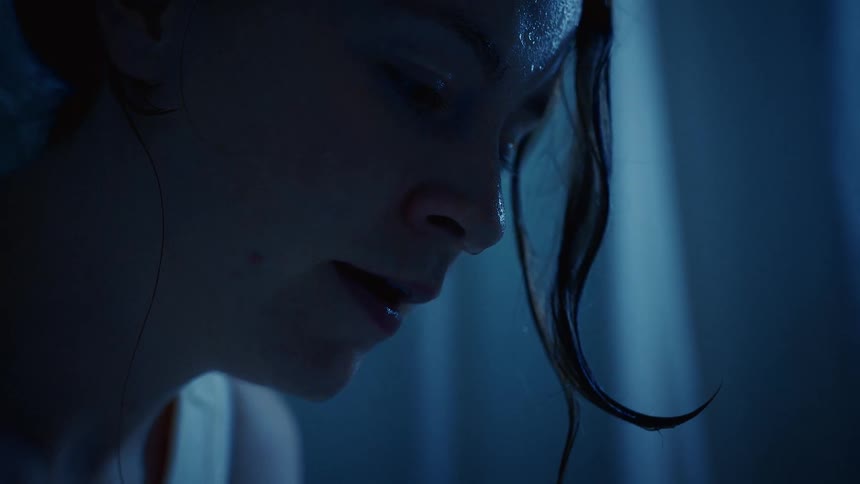 A melancholic woman sits in a dark, blue-lit room as water drips from her wet hair and face in slow motion