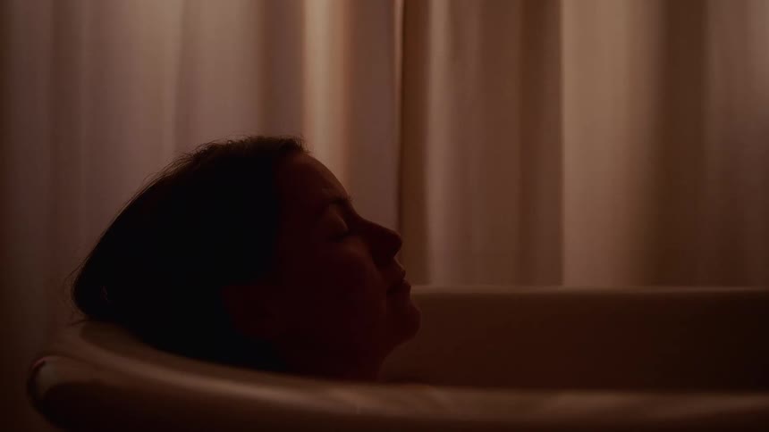 A woman rests her head back and relaxes in a bathtub in a dimly lit, tranquil bathroom
