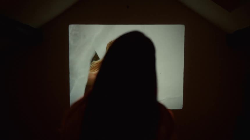 A person sits in silhouette watching a nostalgic vintage home movie projected onto the wall of a dark room