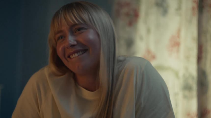 A close-up of a joyful blonde woman with bangs laughing genuinely in slow motion in a warmly lit room