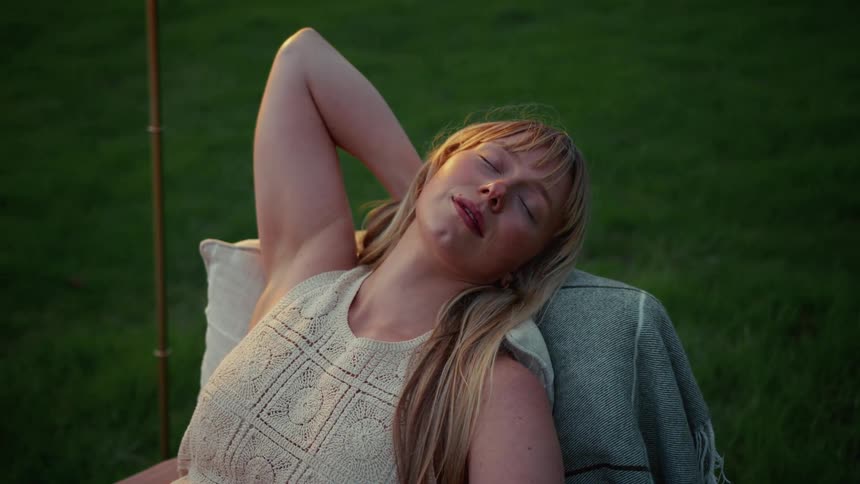 A serene young woman with blonde hair rests in a comfortable chair in a grassy field, waking up peacefully during a beautiful sunset