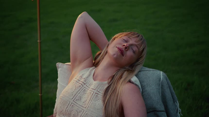 A serene blonde woman rests with her eyes closed in a comfortable chair outdoors at dusk, slowly opening them with a peaceful and contented expression