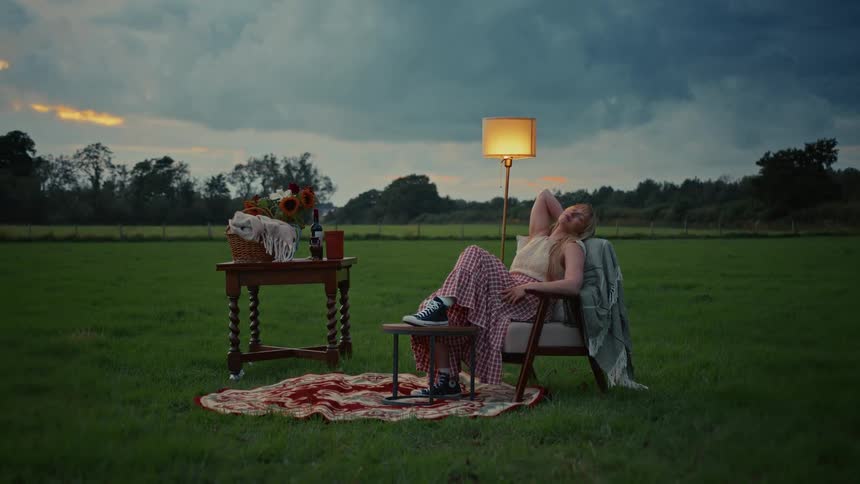 A young woman enjoys a peaceful, surreal moment, lounging in an armchair with a lit lamp in a vast green field at dusk