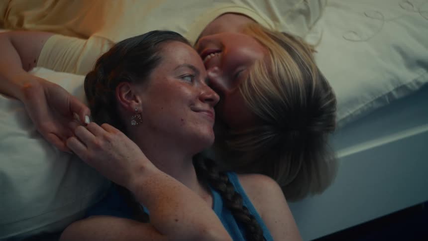 An affectionate female couple smiles and cuddles together in a warmly lit bedroom, sharing an intimate moment