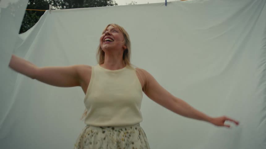 A joyful woman with long blonde hair spins in slow motion with her arms outstretched in front of a white sheet hanging outside
