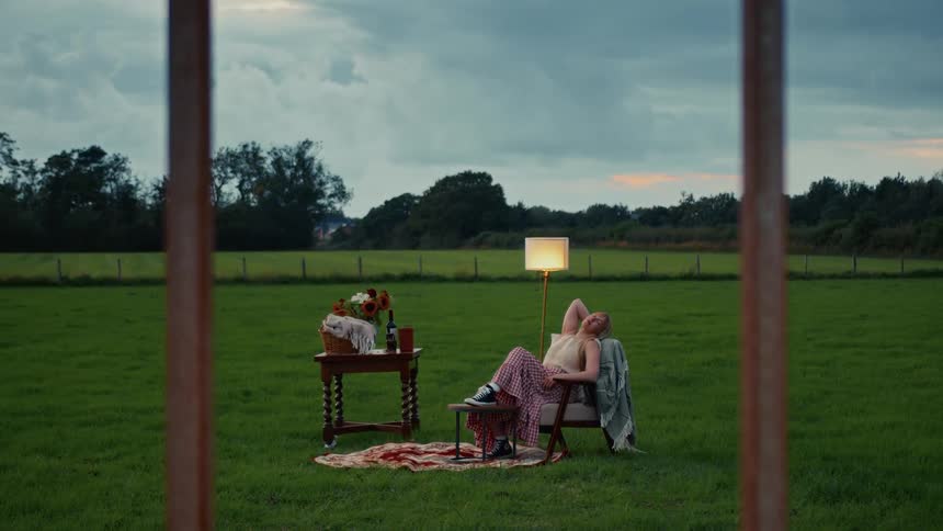 A surreal scene unfolds as a woman relaxes in an armchair with a lamp in a vast green field at dusk, viewed through a window frame