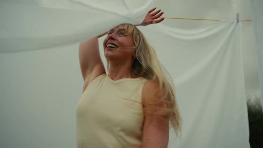 A carefree blonde woman smiles as she peeks out from behind a white sheet swaying in the wind on a clothesline