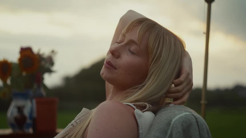A tranquil blonde woman with her eyes closed soaks in the last rays of sun, resting peacefully in a rustic outdoor setting