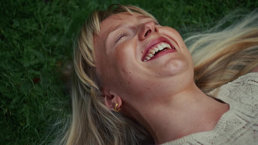 A joyful blonde woman with freckles lies on the green grass, smiling and laughing in a moment of pure bliss